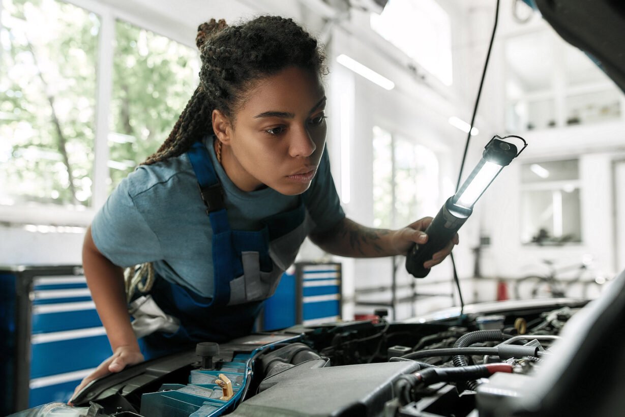 A little care for your vehicle. Young african american woman, professional female mechanic looking, examining under hood of car with torch at auto repair shop