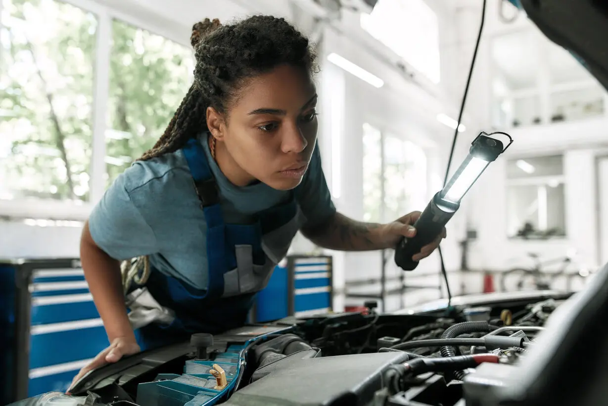 A little care for your vehicle. Young african american woman, professional female mechanic looking, examining under hood of car with torch at auto repair shop